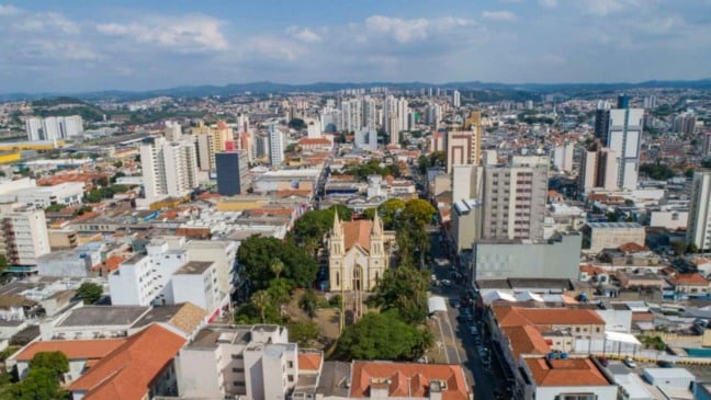 Vista aérea do centro de Jundiaí com prédios, ruas movimentadas e igreja histórica em destaque, cercada por árvores.