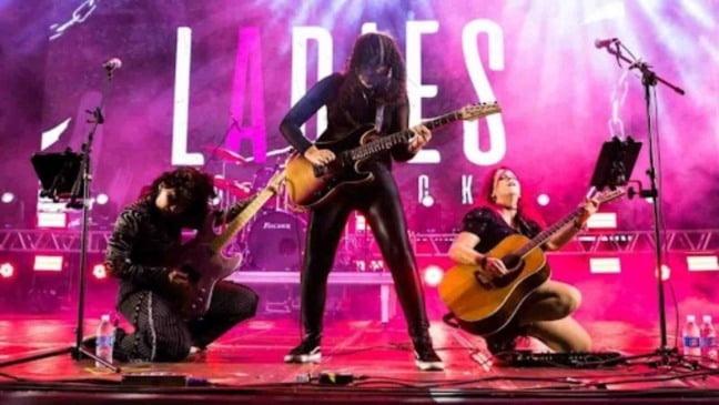Banda feminina Ladies of Rock se apresenta ao vivo no palco, com três musicistas tocando guitarras e violão sob iluminação roxa e rosa, em show de rock com forte presença cênica.