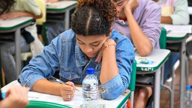Estudante realiza prova em sala de aula, concentrada enquanto escreve respostas, com outros alunos ao fundo.