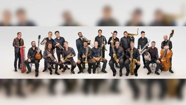 Integrantes da Speakin’ Jazz Big Band posam juntos com instrumentos musicais em foto promocional, destacando saxofones, trompetes, trombones, contrabaixo e acordeão.