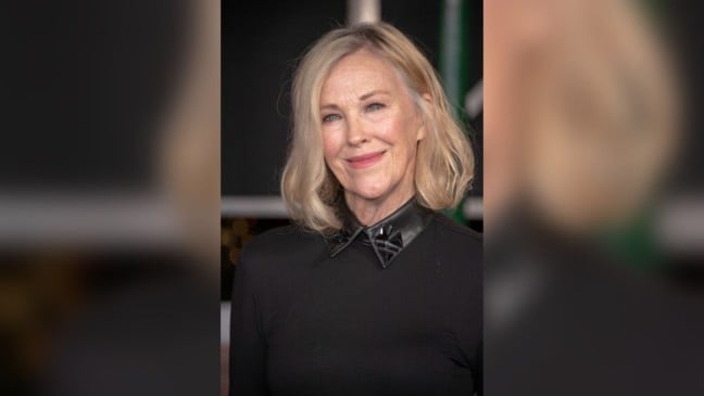 Mulher loira de cabelos curtos e ondulados, vestindo roupa preta com detalhe na gola, sorri para a câmera em evento público.