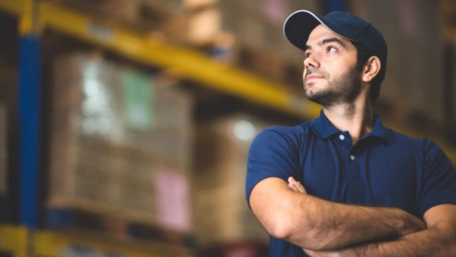 Homem com boné e camiseta azul, de braços cruzados, observa o interior de um galpão com prateleiras e caixas ao fundo.