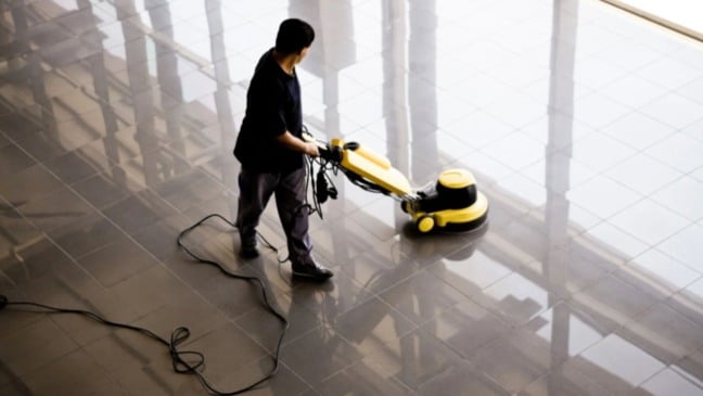Auxiliar de limpeza utiliza equipamento industrial para higienização de piso em área interna de grande circulação.