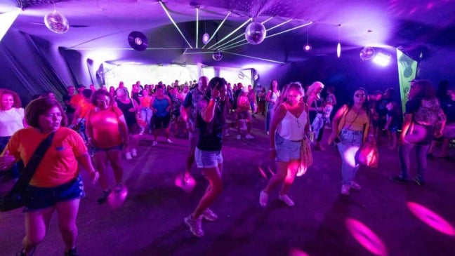 Pessoas de várias idades dançam sob luzes coloridas no Espaço Discoteca da Festa da Uva de Jundiaí.