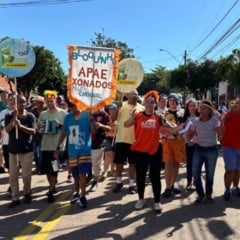 Pessoas caminham em desfile do Bloco APAExonados pelo Carnaval, da APAE de Jundia&iacute;, usando fantasias coloridas e segurando banner do bloco em rua do bairro.