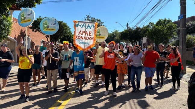 Pessoas caminham em desfile do Bloco APAExonados pelo Carnaval, da APAE de Jundiaí, usando fantasias coloridas e segurando banner do bloco em rua do bairro.