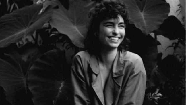 Bruna Caram sorri em retrato em preto e branco, sentada diante de folhas grandes, em clima intimista e artístico.