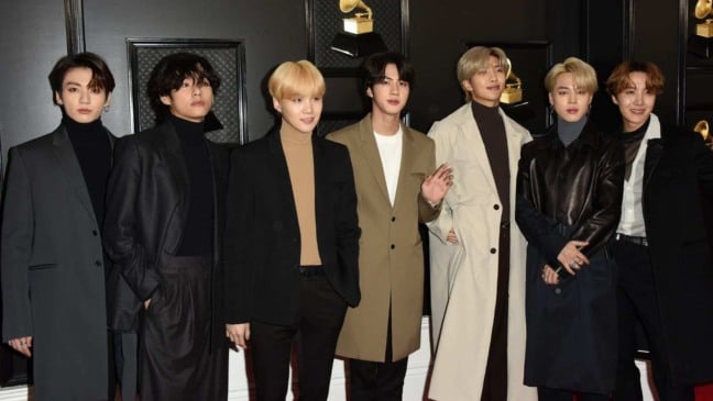 Integrantes do BTS posam juntos em evento de premiação musical, usando ternos e casacos elegantes em tons neutros, diante de um fundo escuro com estatuetas douradas.