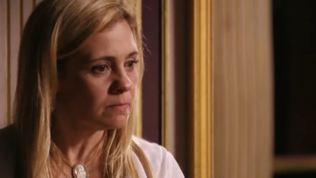 Mulher loira, com expressão séria e olhar fixo, aparece em close dentro de um ambiente interno sofisticado, com molduras douradas ao fundo, transmitindo tensão e intensidade emocional.