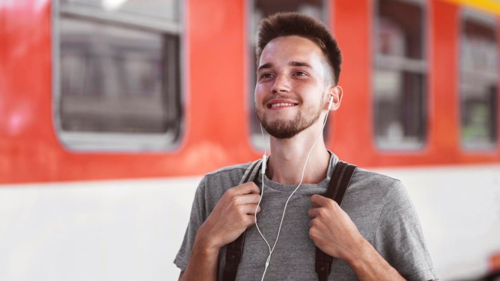 Jovem com mochila e fones de ouvido sorri em plataforma de estação de trem, com composição urbana ao fundo. Jovem com mochila e fones de ouvido sorri em plataforma de estação de trem, com composição urbana ao fundo.