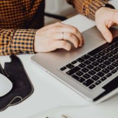 Pessoa digitando em notebook em ambiente de trabalho, com mouse sem fio e x&iacute;cara de caf&eacute; sobre a mesa.