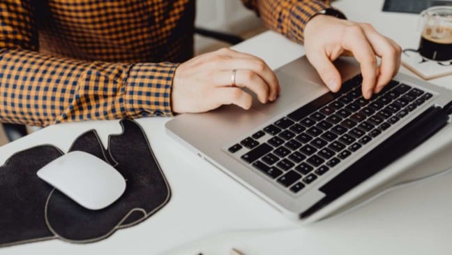 Pessoa digitando em notebook em ambiente de trabalho, com mouse sem fio e xícara de café sobre a mesa.