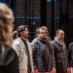 Grupo de homens ensaia canto coral em palco, acompanhando a regente durante prepara&ccedil;&atilde;o musical.