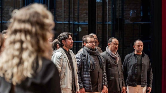 Grupo de homens ensaia canto coral em palco, acompanhando a regente durante preparação musical.