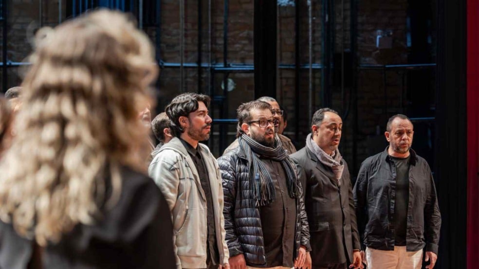 Grupo de homens ensaia canto coral em palco, acompanhando a regente durante preparação musical. Grupo de homens ensaia canto coral em palco, acompanhando a regente durante preparação musical.
