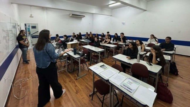 Professora e educador conduzem aula em sala do Cursinho Popular Chico Poço, com estudantes sentados em carteiras, materiais de estudo sobre as mesas e ambiente voltado à preparação para vestibulares.