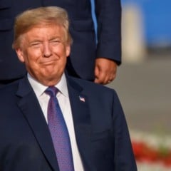 Trump sorrindo durante evento oficial ao ar livre, vestindo terno azul e gravata vermelha