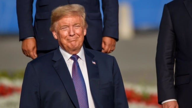 Trump sorrindo durante evento oficial ao ar livre, vestindo terno azul e gravata vermelha