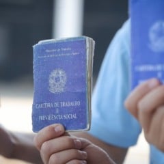 Pessoas seguram carteiras de trabalho azuis, documento usado para registro de emprego formal no Brasil.