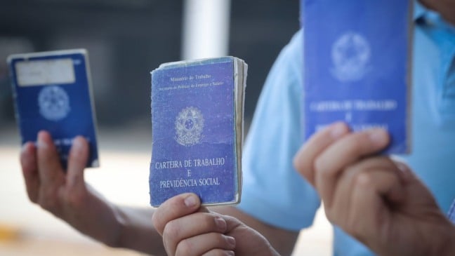 Pessoas seguram carteiras de trabalho azuis, documento usado para registro de emprego formal no Brasil.
