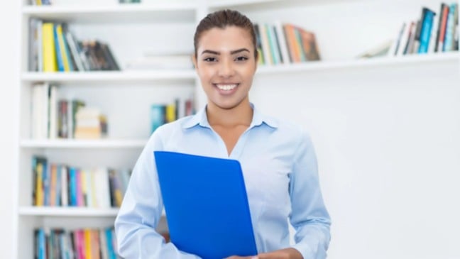 Jovem mulher sorridente segura uma pasta azul em ambiente com estantes de livros, representando estudante ou estagiária em contexto educacional e profissional.