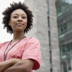 Profissional da &aacute;rea da sa&uacute;de de uniforme rosa posa com bra&ccedil;os cruzados em frente a pr&eacute;dio institucional, transmitindo confian&ccedil;a e profissionalismo.