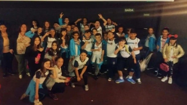 Grupo de alunos da rede municipal de Jundiaí, em uniforme escolar, posa animado em espaço interno, acompanhado por professores.