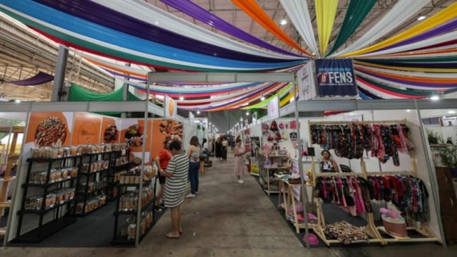 Corredor da FENS em Jundiaí com estandes de empreendedores, pessoas circulando e decoração colorida no teto do pavilhão.