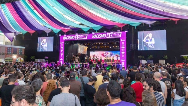 Público acompanha apresentação no palco principal da Festa da Uva de Jundiaí, com decoração colorida e mensagem “Racismo é crime” em destaque.