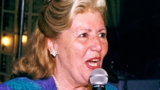 Florência Passarin fala ao microfone durante evento social, vestindo roupa azul e roxa, com expressão confiante e elegante.