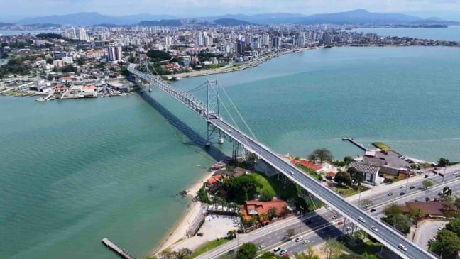 Vista aérea da Ponte Hercílio Luz, em Florianópolis, ligando a ilha ao continente, com o mar, prédios e vias urbanas ao redor.