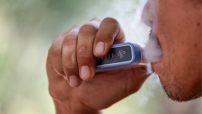 Pessoa utiliza cigarro eletrônico e solta vapor, representando o uso de vape e a dependência de nicotina.