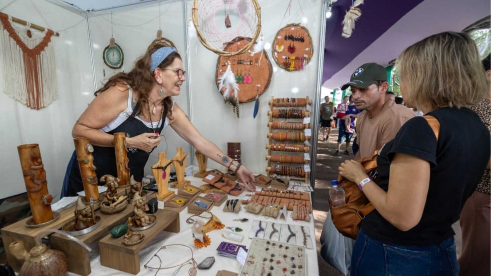 Artesã apresenta acessórios e objetos artesanais em estande da Festa da Uva, enquanto visitantes observam e escolhem produtos feitos com materiais naturais. Artesã apresenta acessórios e objetos artesanais em estande da Festa da Uva, enquanto visitantes observam e escolhem produtos feitos com materiais naturais.