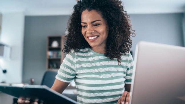 Mulher sorrindo enquanto trabalha em home office, usando notebook e tablet em ambiente interno.