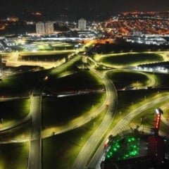 Vista a&eacute;rea noturna de rodovias com trevos e al&ccedil;as iluminados por LED, mostrando ilumina&ccedil;&atilde;o uniforme, tr&aacute;fego leve e a cidade ao fundo.