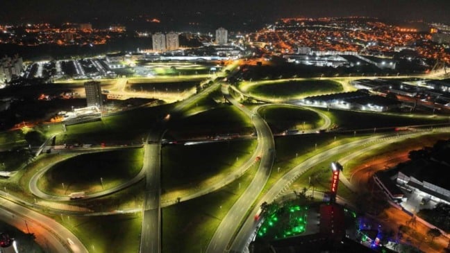 Vista aérea noturna de rodovias com trevos e alças iluminados por LED, mostrando iluminação uniforme, tráfego leve e a cidade ao fundo.