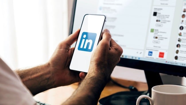 Pessoa segurando um celular com o aplicativo do LinkedIn aberto, enquanto a rede social aparece ao fundo na tela de um computador em ambiente de trabalho.