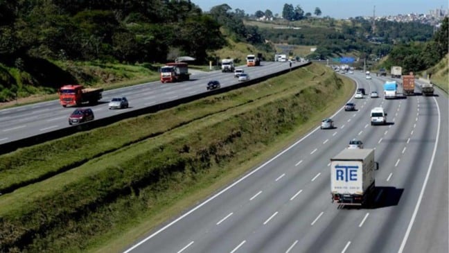 Rodovia do Sistema Anhanguera-Bandeirantes com tráfego intenso de caminhões e carros, pistas duplicadas e canteiro central verde.