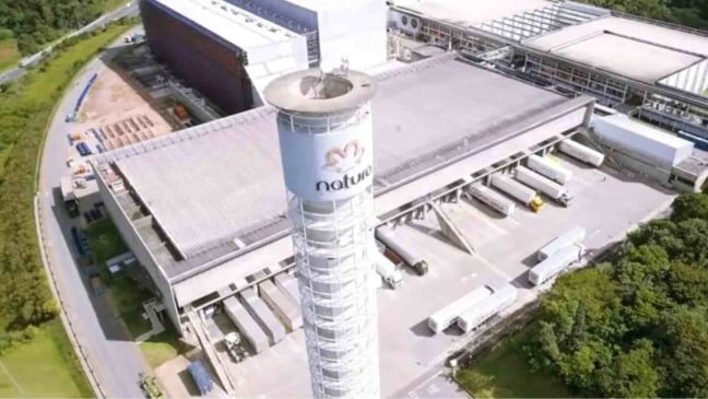 Vista aérea do complexo industrial da Natura, com torre central com o logotipo da empresa, galpões, caminhões e área verde ao redor.