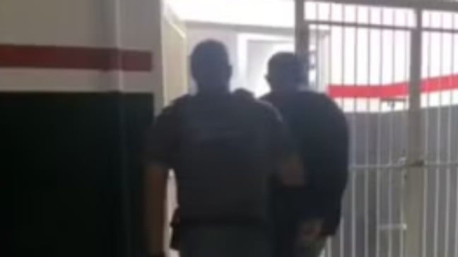 Policiais escoltam homem detido por corredor interno de delegacia em direção a área com grades metálicas.