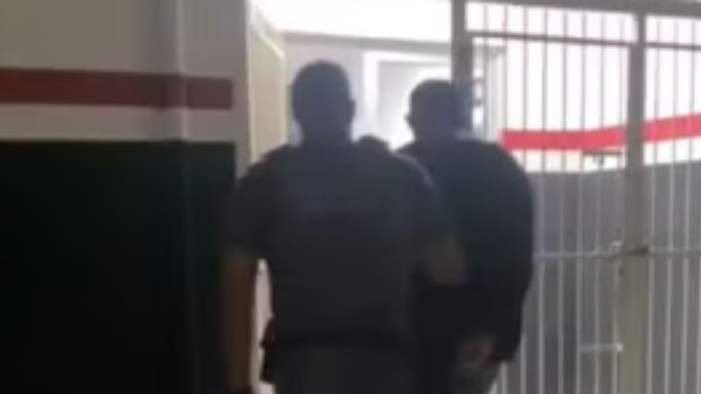 Policiais escoltam homem detido por corredor interno de delegacia em direção a área com grades metálicas. Policiais escoltam homem detido por corredor interno de delegacia em direção a área com grades metálicas.