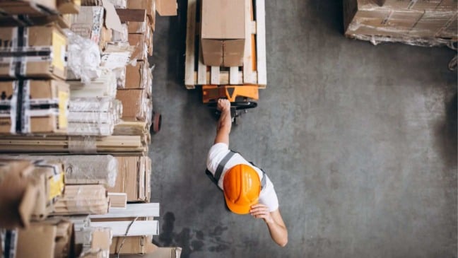 Trabalhador com capacete manuseia paleteira em almoxarifado, movimentando caixas empilhadas em um galpão de armazenagem.