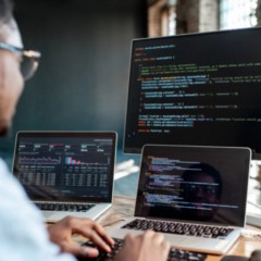 Profissional de tecnologia trabalhando com programa&ccedil;&atilde;o em notebooks e monitor, exibindo c&oacute;digos e gr&aacute;ficos em ambiente de escrit&oacute;rio moderno.