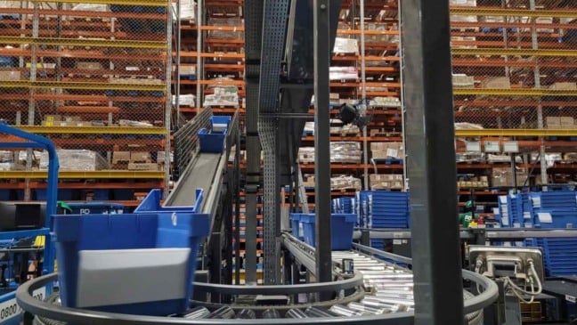 Interior de centro de distribuição com esteiras automáticas, caixas azuis e prateleiras industriais usadas na operação logística.