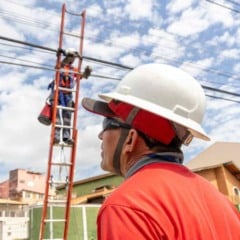 Trabalhadores realizam reordenamento de cabos em postes de rua residencial, com t&eacute;cnico em escada organizando a fia&ccedil;&atilde;o a&eacute;rea e outro acompanhando o servi&ccedil;o no solo.