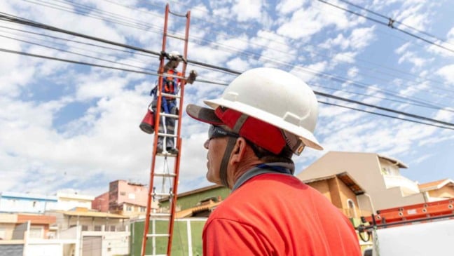 Trabalhadores realizam reordenamento de cabos em postes de rua residencial, com técnico em escada organizando a fiação aérea e outro acompanhando o serviço no solo.