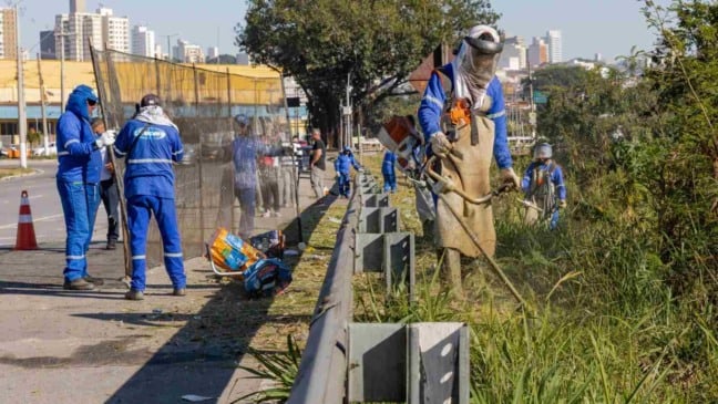 Equipes da zeladoria urbana realizam roçagem e limpeza às margens de avenida em Jundiaí, com uso de equipamentos de proteção e máquinas de corte de vegetação.