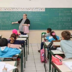 Professora ministra aula para alunos do ensino fundamental em sala de aula da rede municipal, com crian&ccedil;as sentadas em carteiras e quadro-negro ao fundo.