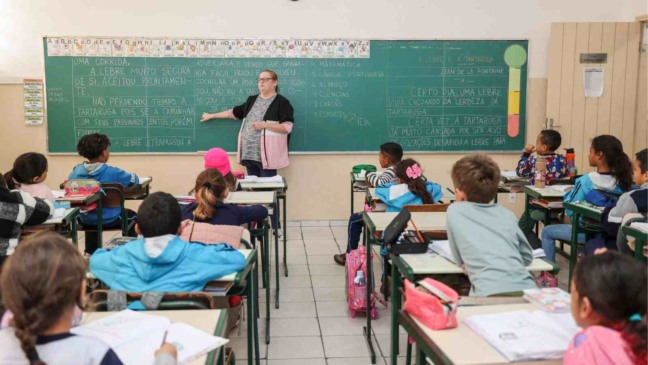 Professora ministra aula para alunos do ensino fundamental em sala de aula da rede municipal, com crianças sentadas em carteiras e quadro-negro ao fundo.