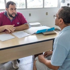 Profissional de sa&uacute;de realiza atendimento a paciente durante consulta em unidade de sa&uacute;de de Jundia&iacute;, com prontu&aacute;rios sobre a mesa e ambiente cl&iacute;nico ao fundo.